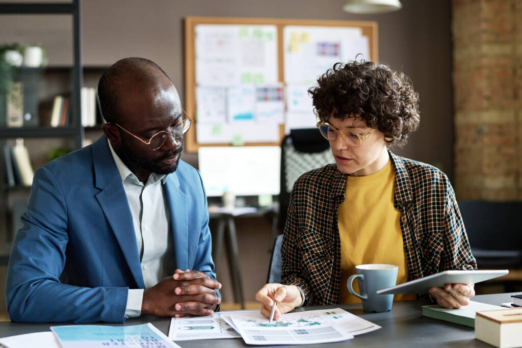 Two professionals in an office setting reviewing financial documents and charts. A woman holds a tablet and gestures toward the papers while discussing strategy with a man in a blue blazer, representing Centura Advisors’ collaborative financial planning approach.